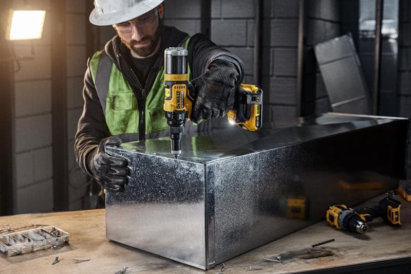 Egy személy védőfelszerelésben DEWALT DCF403NT akkumulátoros elektromos szerszámmal dolgozik egy fém dobozon egy ipari műhelyben. Különböző csavarok és szerszámok láthatók a munkapadon.