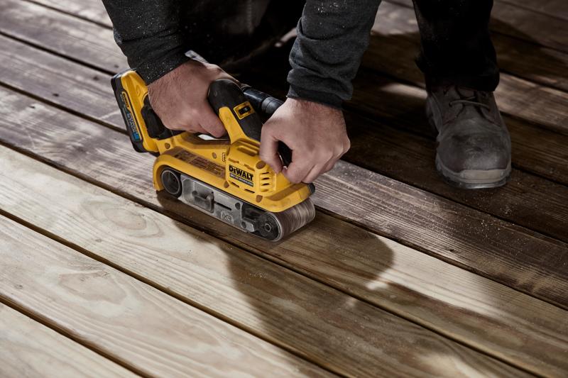 Egy személy sárga DEWALT vezeték nélküli szalagcsiszolót használ fa deszkák felületének simítására.